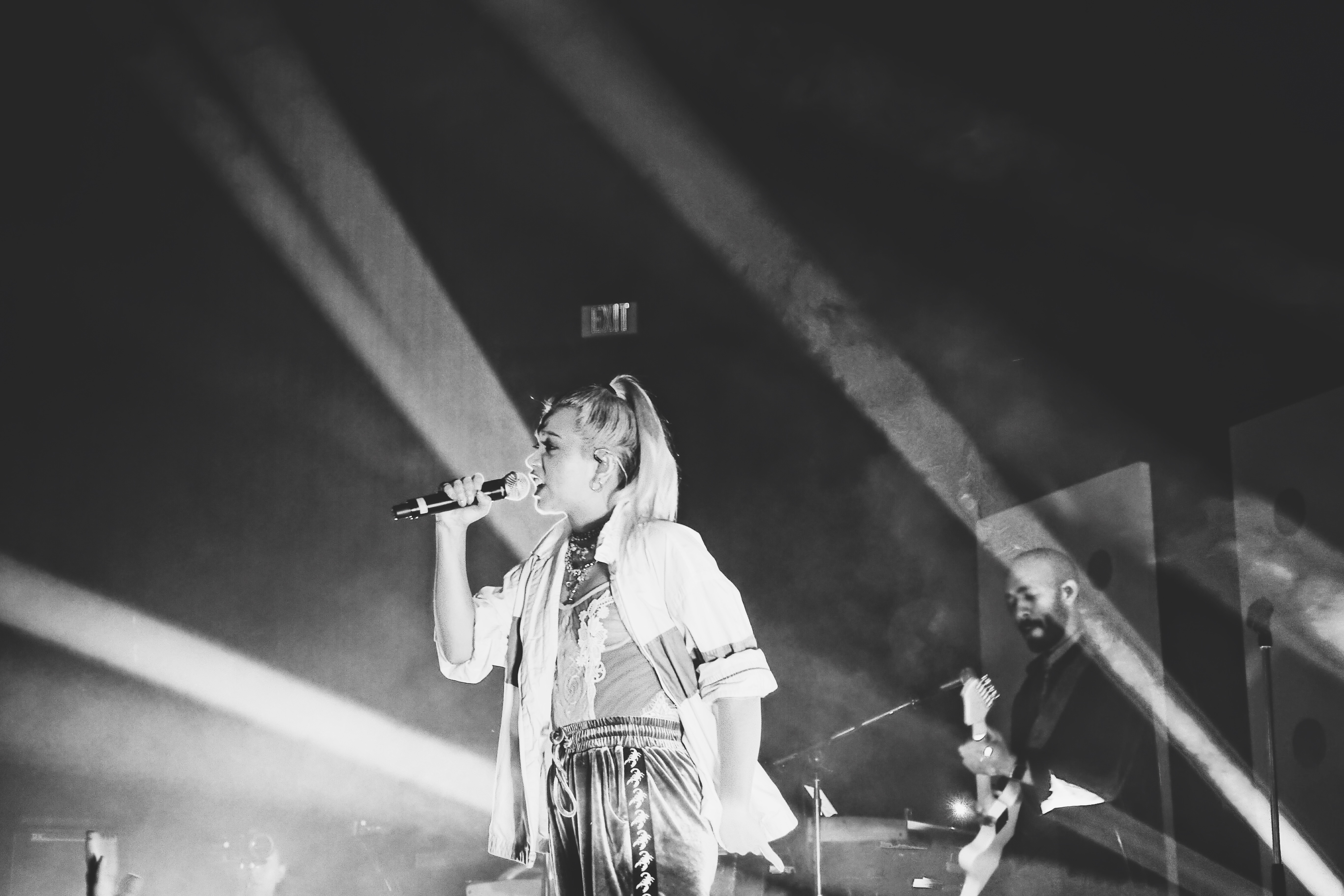 Musician Hayley Kiyoko in black and white, singing on a stage with lights behind her. The stage is filled with smoke from dry ice. An EXIT sign looms above her head in the distance. Her bass player leans into their instrument behind her.